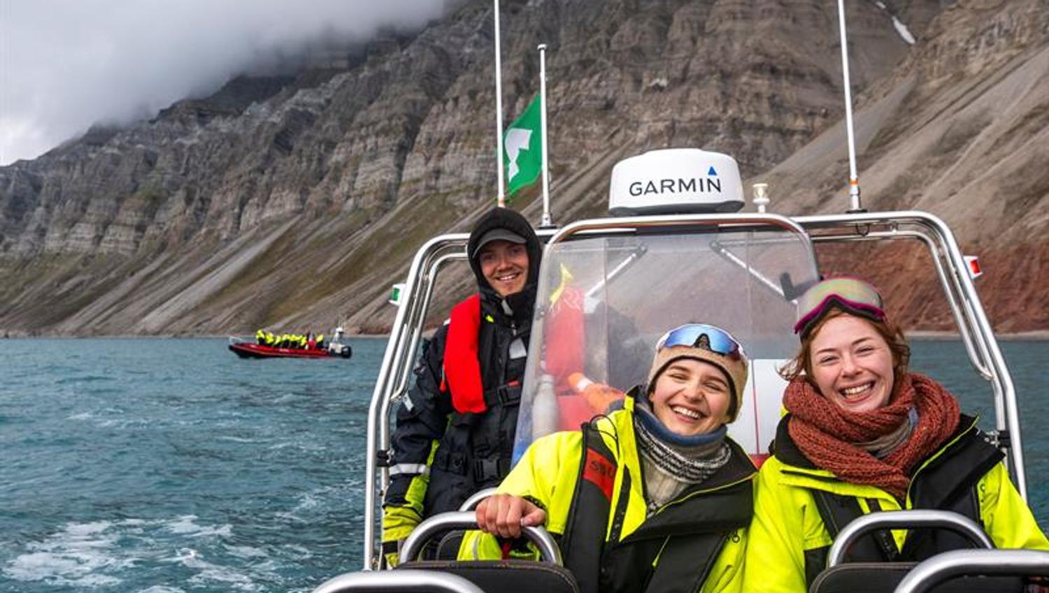 Tre personer smiler på RIB-båt med fjell i bakgrunnen.