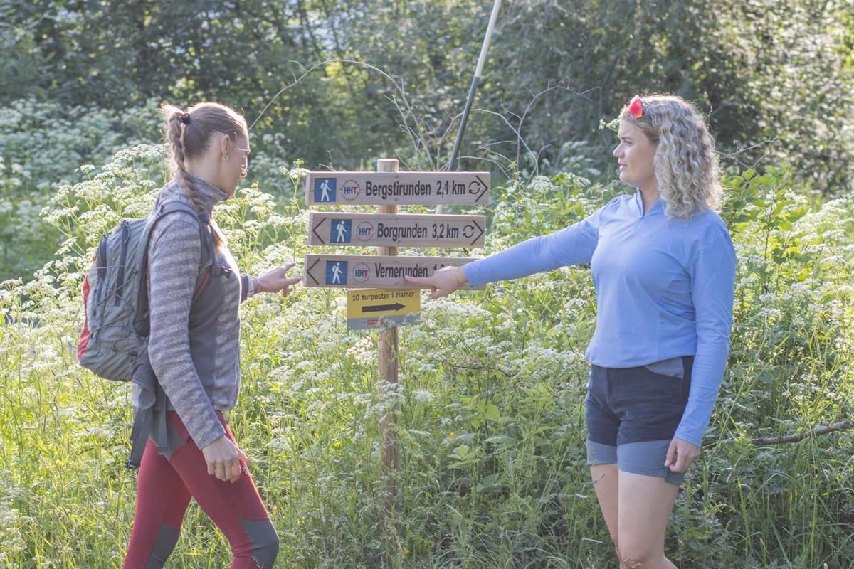 To personer står ved et skilt i en grønn skog, og peker på turstier merket med avstand.