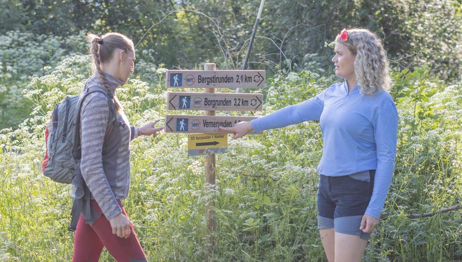 To personer står ved et skilt i en grønn skog, og peker på turstier merket med avstand.