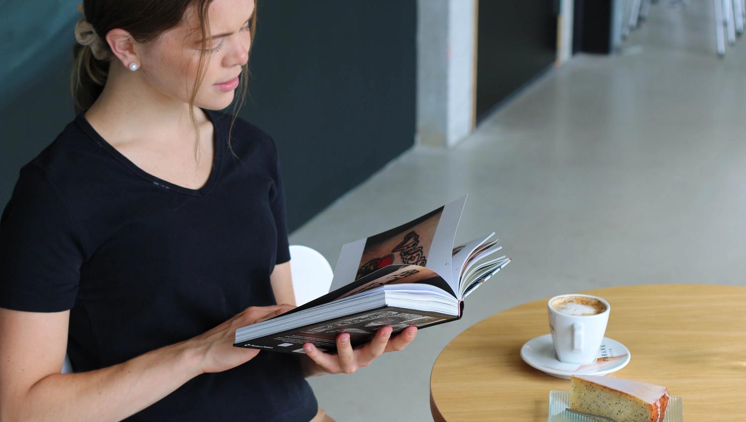 Ei ung kvinne sit ved eit bord og leser i ein bok, med ein kopp kaffi og eit kakestykke foran seg.