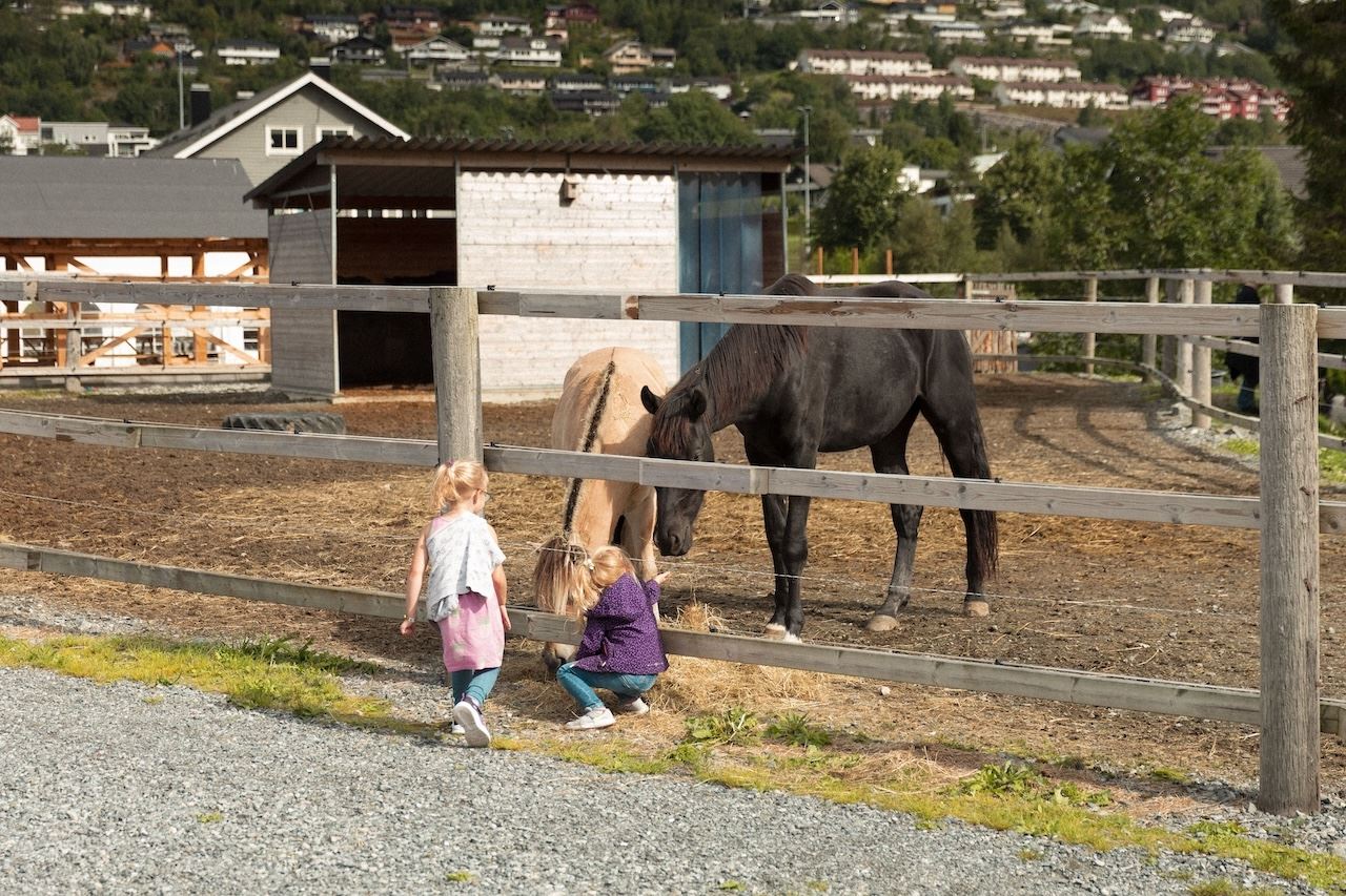To små barn ved eit tregjerde ser på og klappar to hestar i ein innhegning på ein gard, med hus i bakgrunnen..