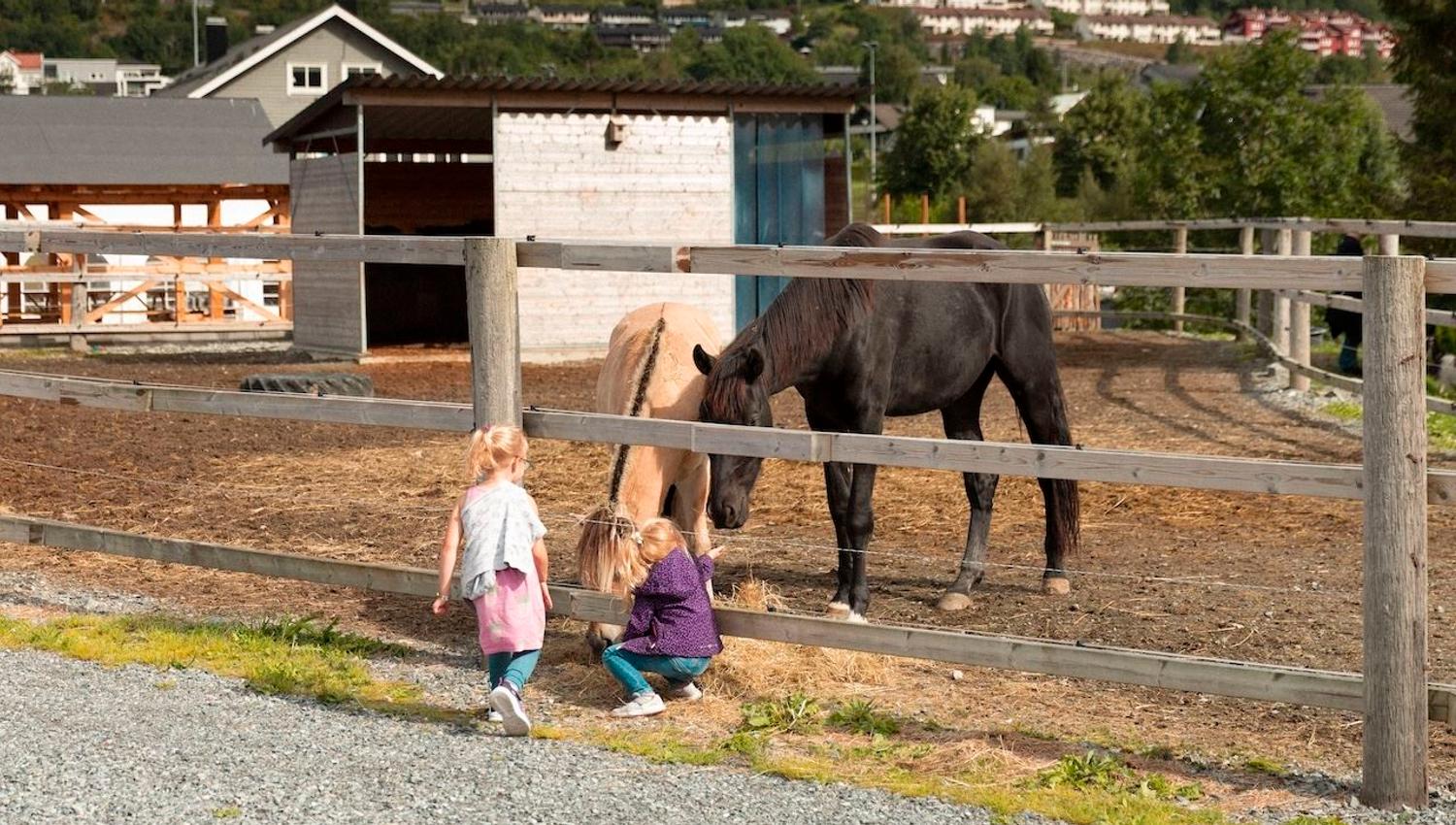 To små barn ved eit tregjerde ser på og klappar to hestar i ein innhegning på ein gard, med hus i bakgrunnen..