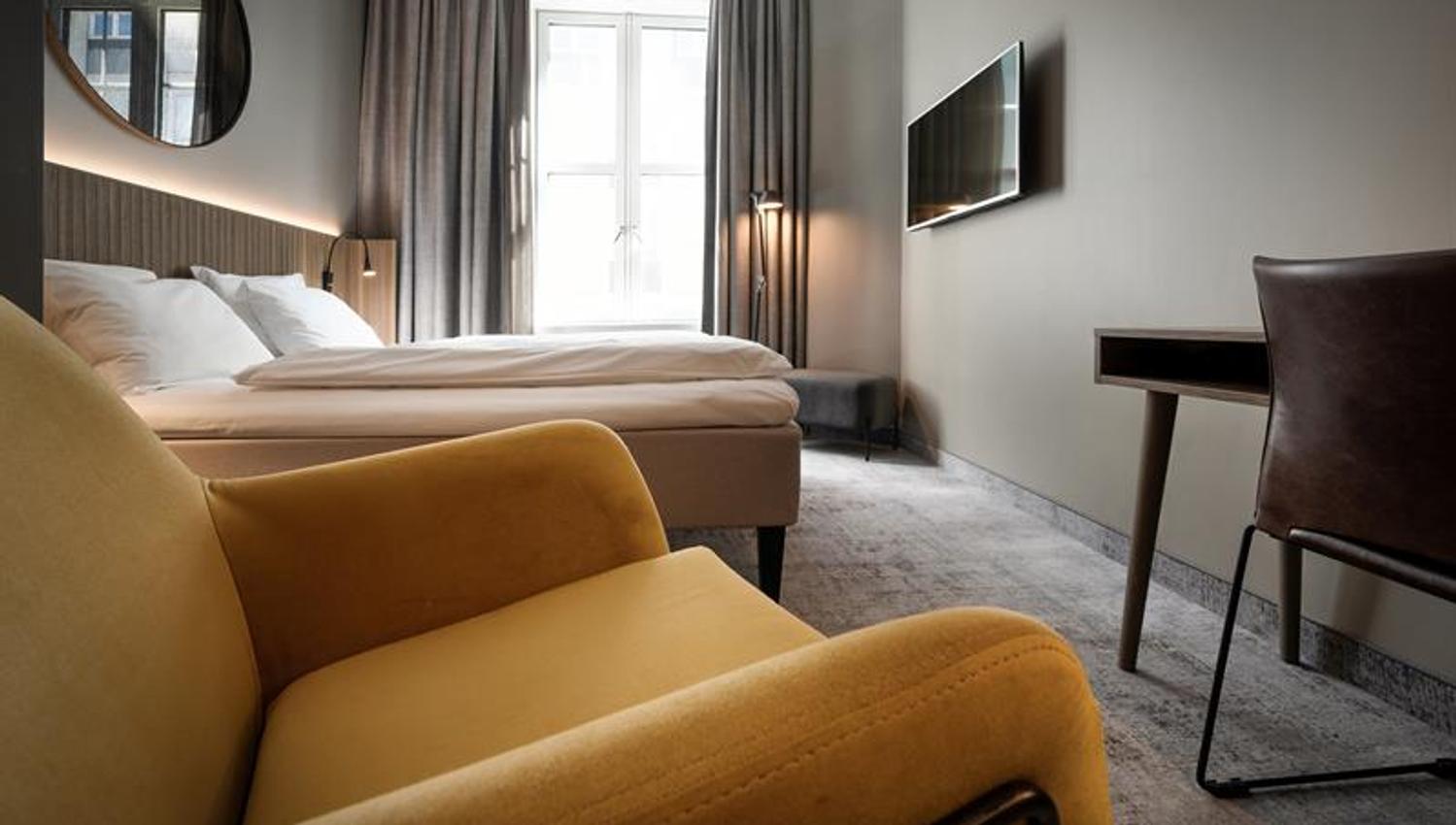 Standard room with a yellow armchair, large double bed, desk and a TV screen on the wall.