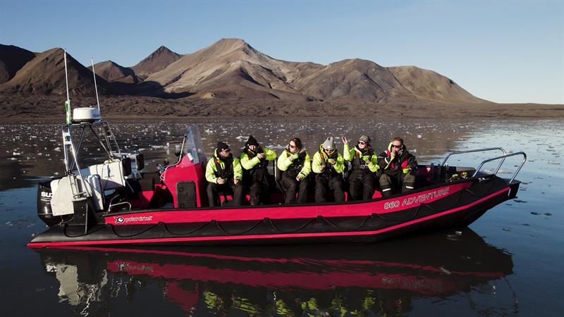 RIB-båt med passasjerer i gule drakter på speilblankt vann med fjell bak.