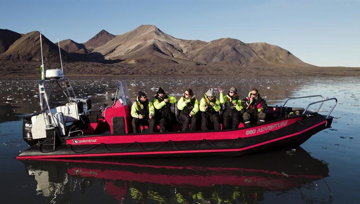 RIB-båt med passasjerer i gule drakter på speilblankt vann med fjell bak.