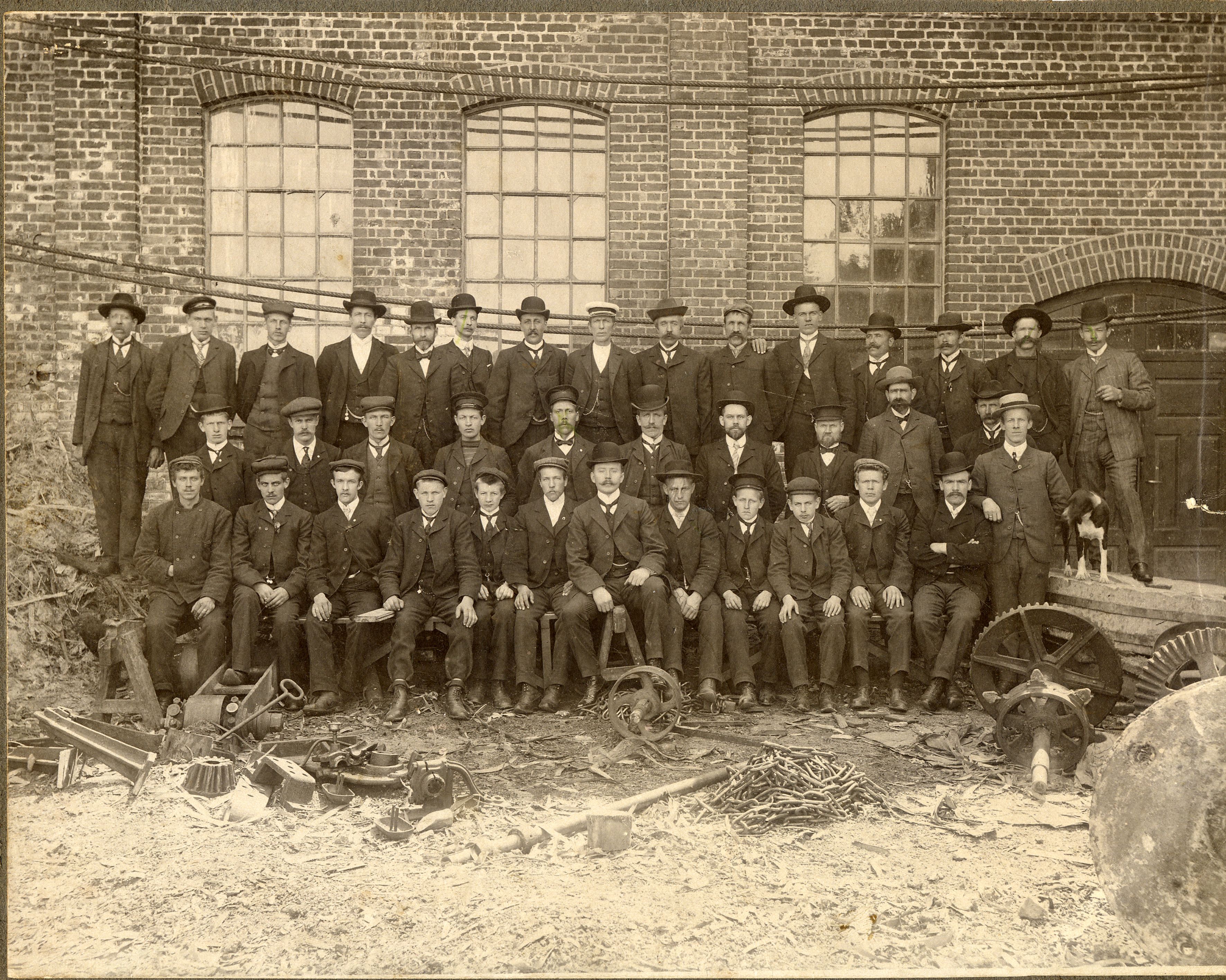 Bildet er et gammelt svart-hvitt foto som viser en stor gruppe menn foran en murbygning. De fleste sitter eller står i rekker og har på seg dresser, hatter og slips. Foran dem ligger metall hjul og verktøy, noe som tyder på at de jobbet på en fabrikk eller et verksted.