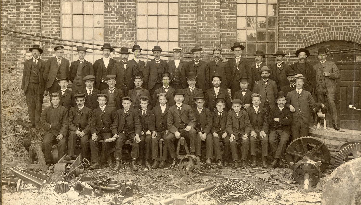 Bildet er et gammelt svart-hvitt foto som viser en stor gruppe menn foran en murbygning. De fleste sitter eller står i rekker og har på seg dresser, hatter og slips. Foran dem ligger metall hjul og verktøy, noe som tyder på at de jobbet på en fabrikk eller et verksted.