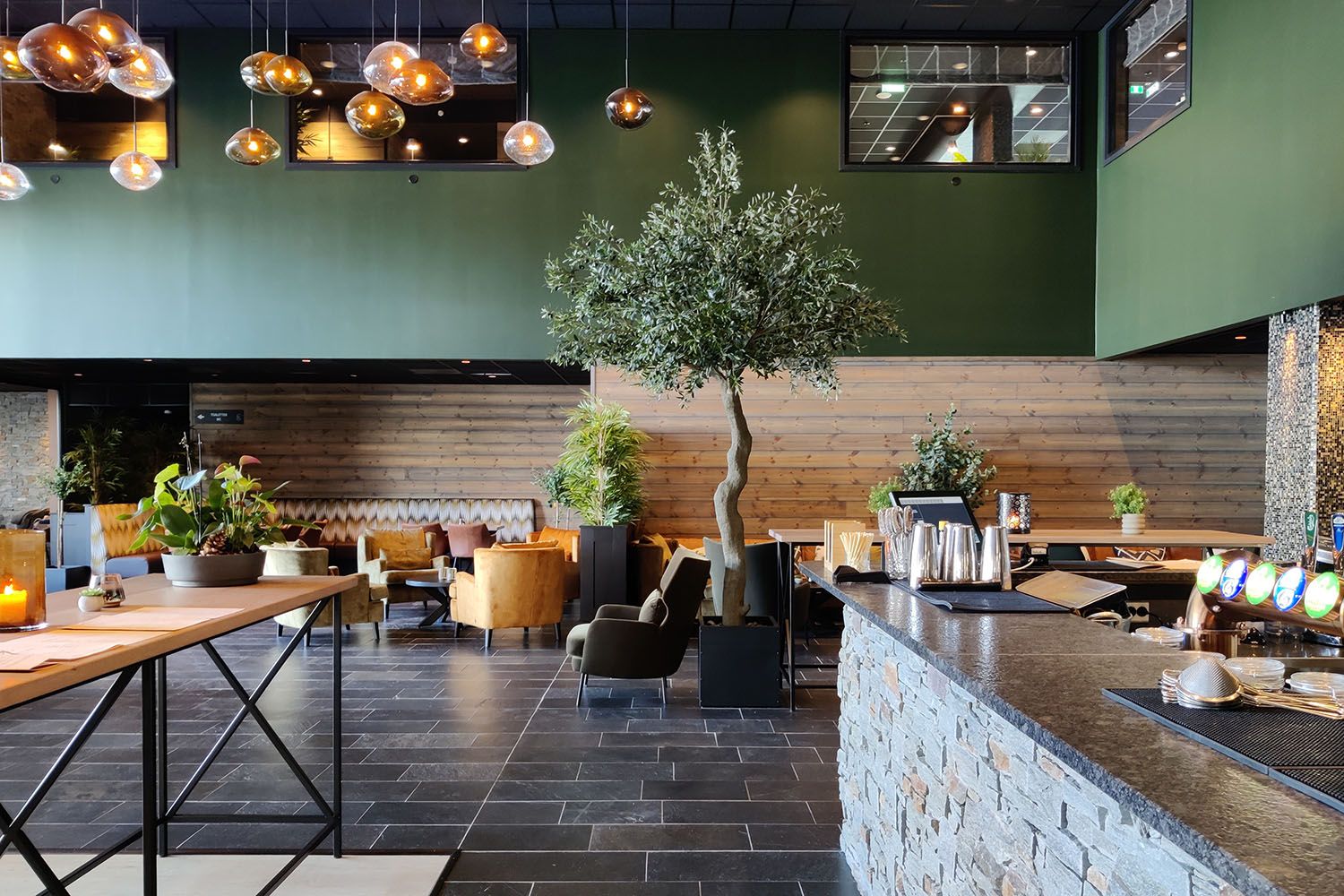 Restaurantinteriør med skifergulv, murt bardisk, trevegg med trehvit liggende panel, grønn forblending og mange lune lamper som henger fra taket. Sentralt står et oliventre.