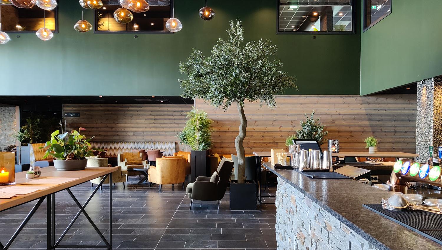 Restaurantinteriør med skifergulv, murt bardisk, trevegg med trehvit liggende panel, grønn forblending og mange lune lamper som henger fra taket. Sentralt står et oliventre.