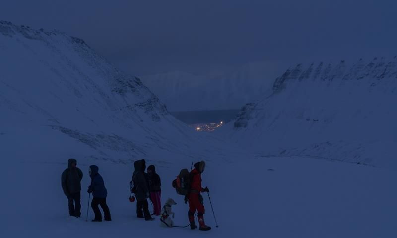 Flere personer og en hund på vei til isgrotten, med Longyearbyen, fjell og fjord i bakgrunnen