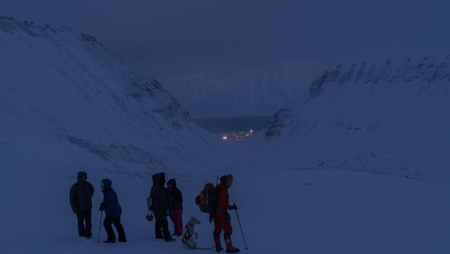 Flere personer og en hund på vei til isgrotten, med Longyearbyen, fjell og fjord i bakgrunnen