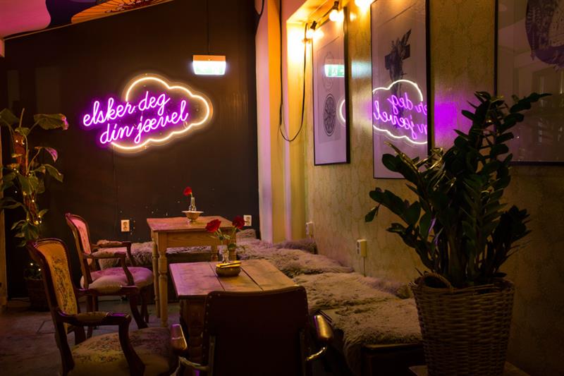 Interior with tables, chairs and a seating sections, as well as plants on each side, as well as a neon sign.