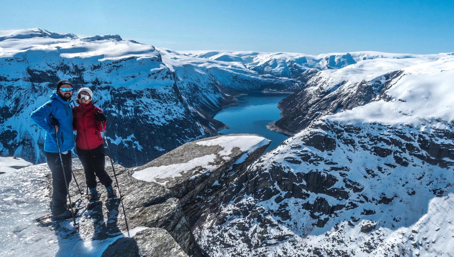 Couple with snowshoes standing on Trolltunga overlooking the icy fjord and mountains.
