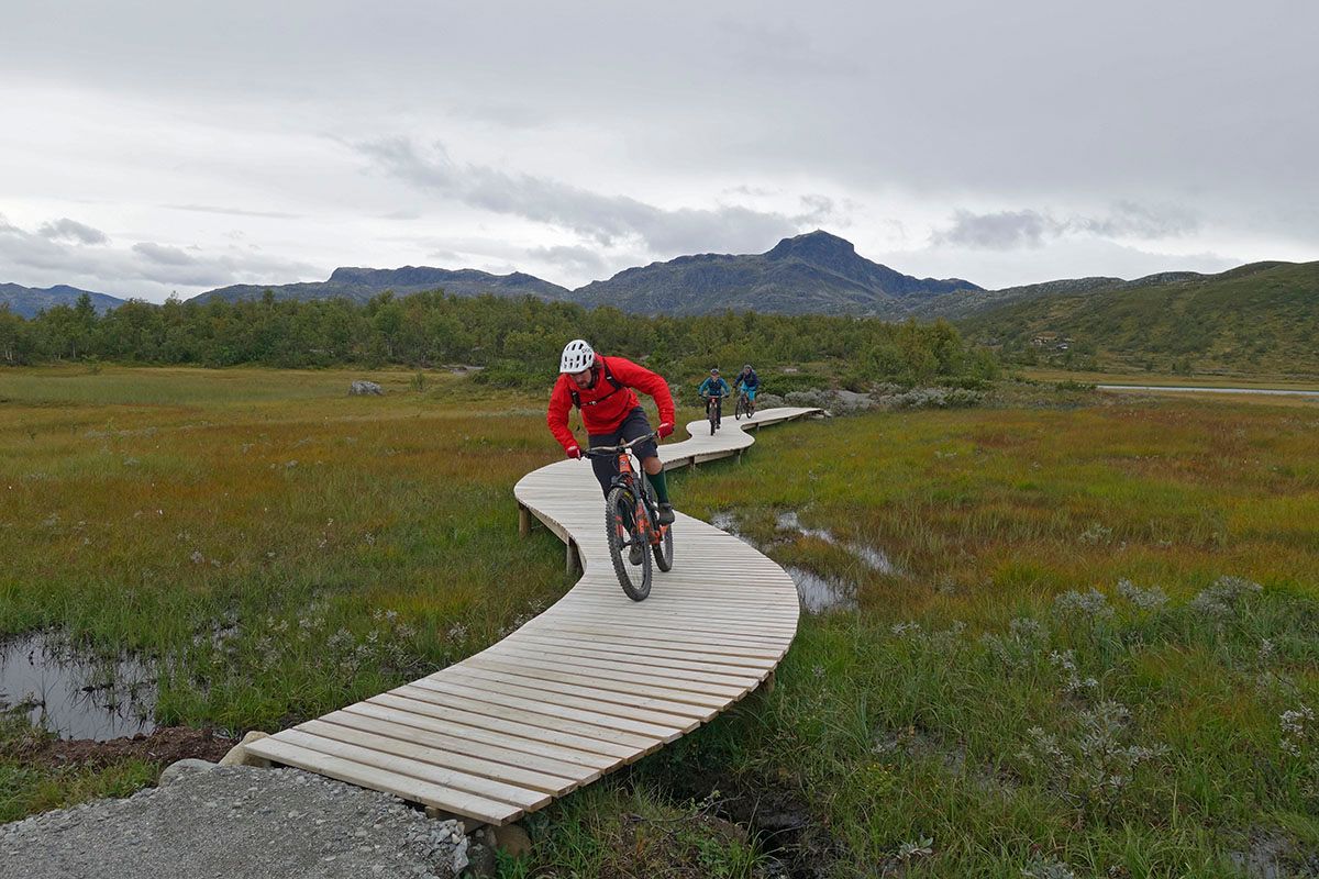 Syklister på bærekraftig bygget terrainsykkelsti med kloppelement over et myr. Fjell i bakgrunnen.