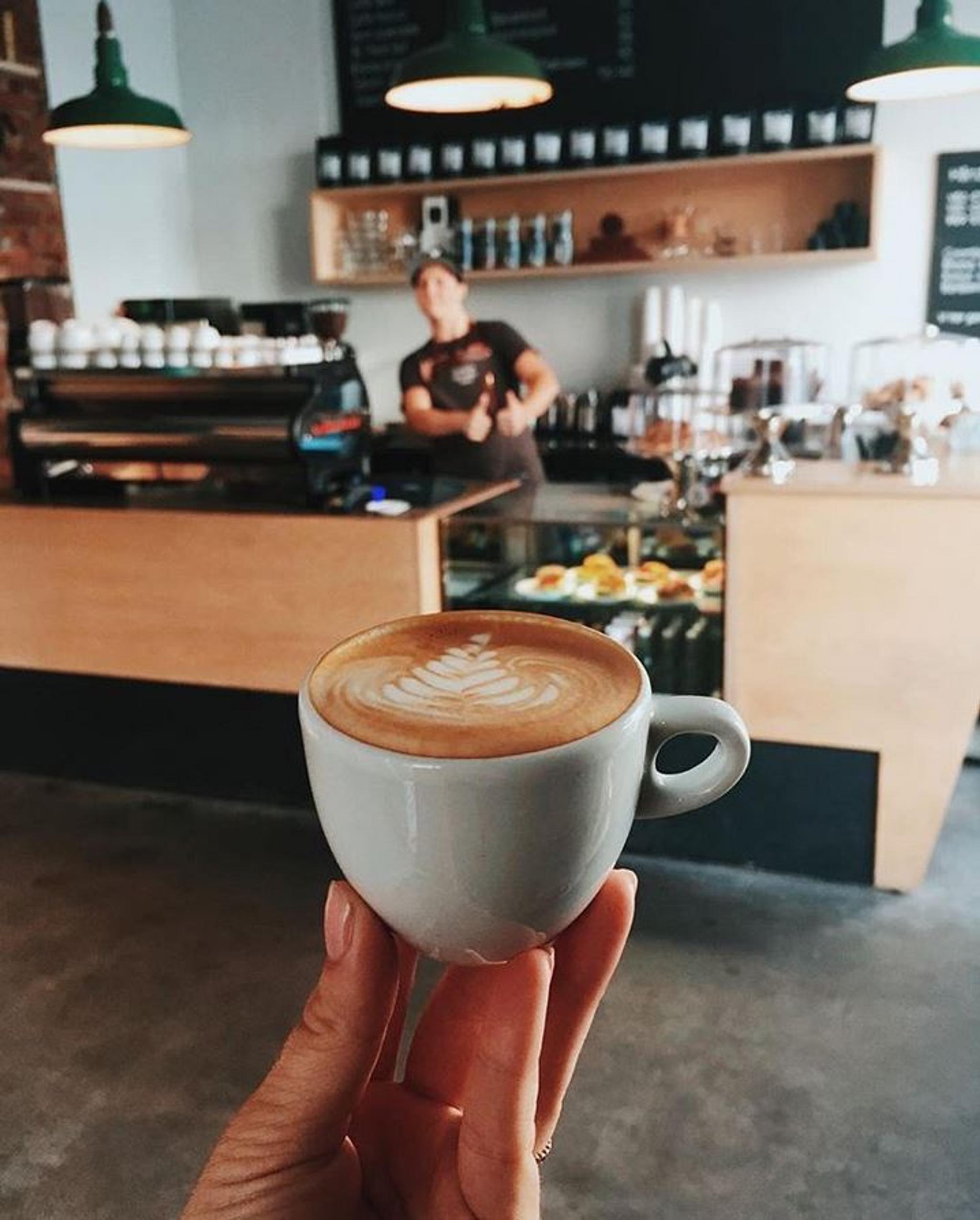 en hånd som viser frem en kaffe med kaffebaren i bakgrunnen