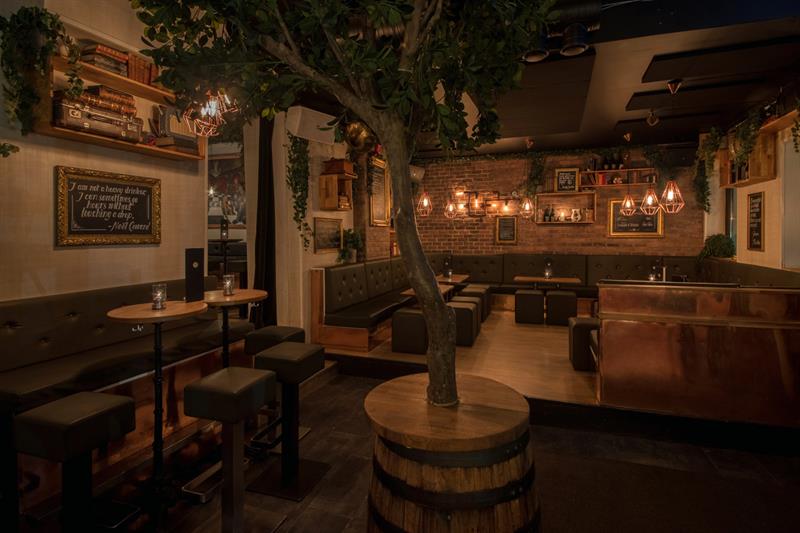 Bar interior with bar counter, bar stools, small tables and chairs, and a tree standing in a barrel in the middle of the room.
