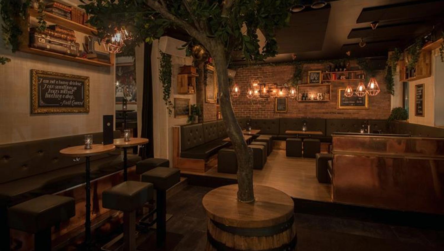 Bar interior with bar counter, bar stools, small tables and chairs, and a tree standing in a barrel in the middle of the room.