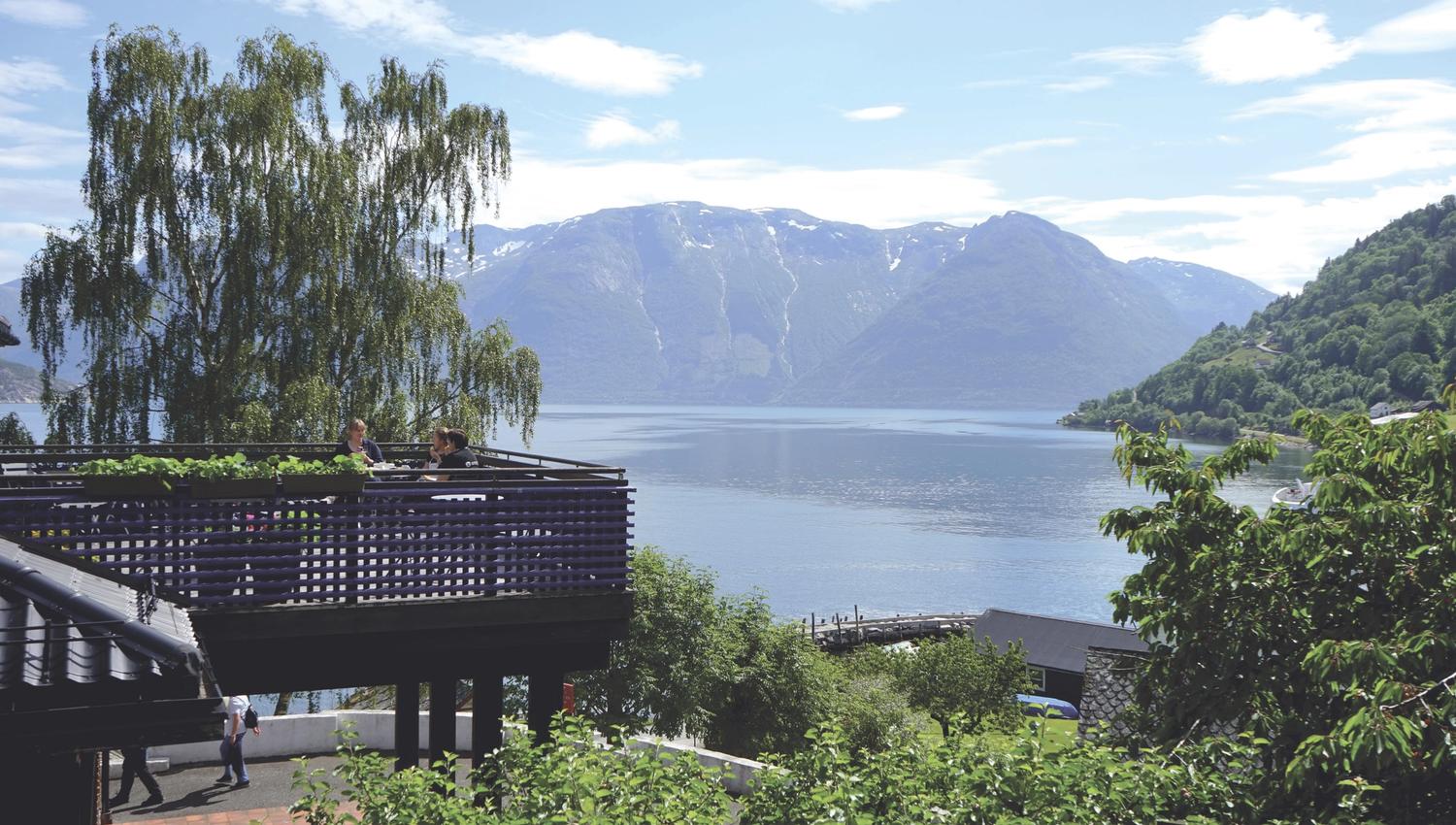 Utsikt ut over fjord og fjell. I forgrunnen er det grønne trær og en altan hvor det sitter tre mennesker.