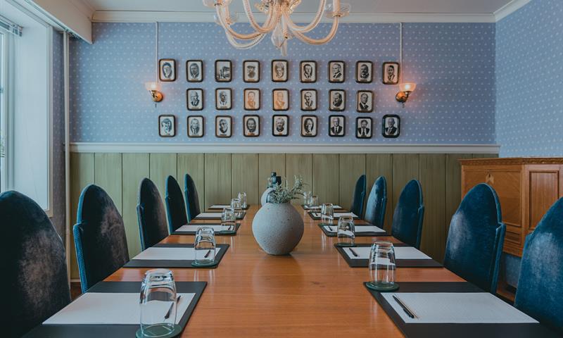 Et møterom med et bord, blå stoler og vegger, med gamle portrettbilder på veggen i bakgrunnen