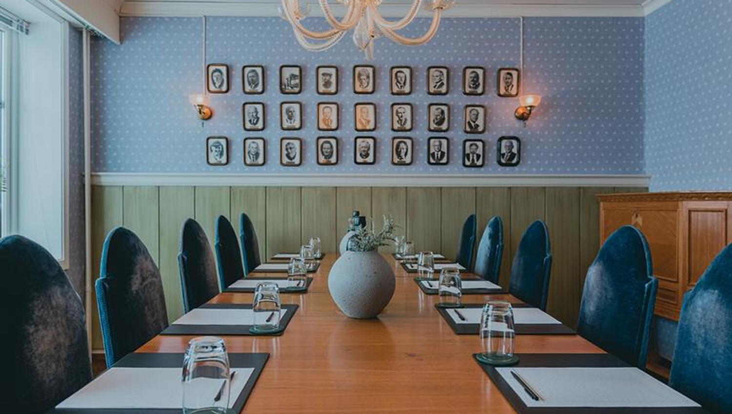 Et møterom med et bord, blå stoler og vegger, med gamle portrettbilder på veggen i bakgrunnen