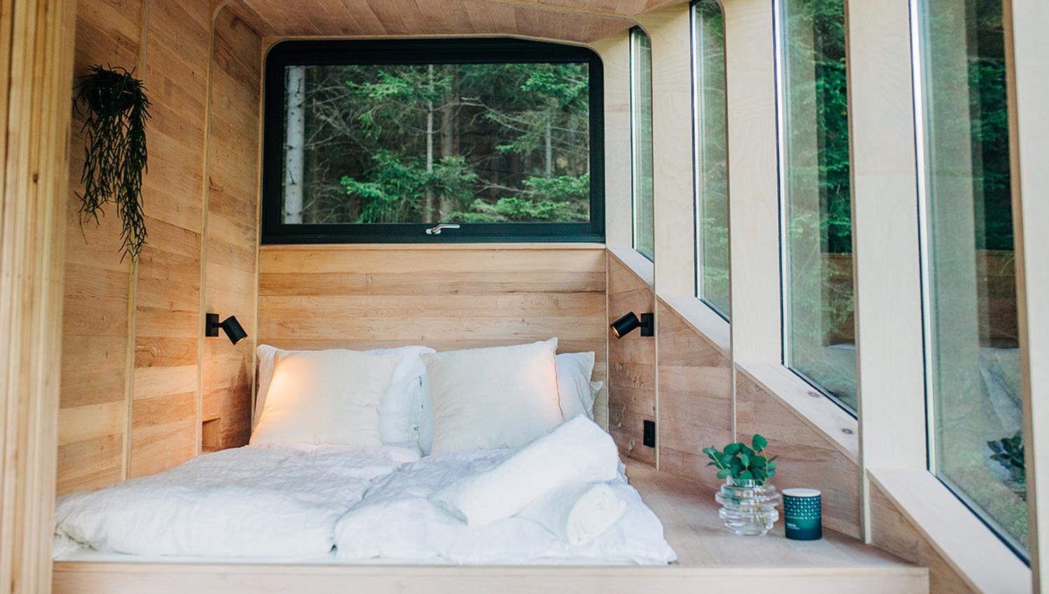 Ei seng med mjuke tekstilar og store vindauge som slepp inn naturlyset i Woodnest tretopphytte.