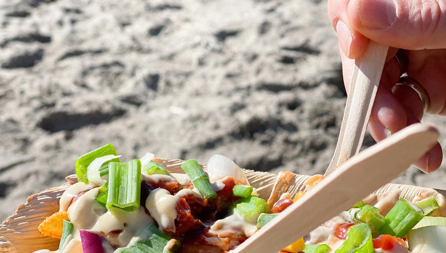 En hånd som holder i en tallerken loaded fries . Sandstrand i bakgrunnen. Foto.