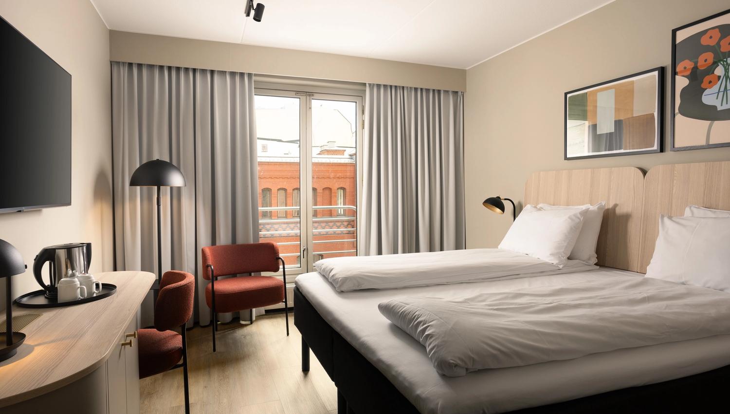 Hotel room with large double bed, TV on the wall, red chair in the corner and grey curtains