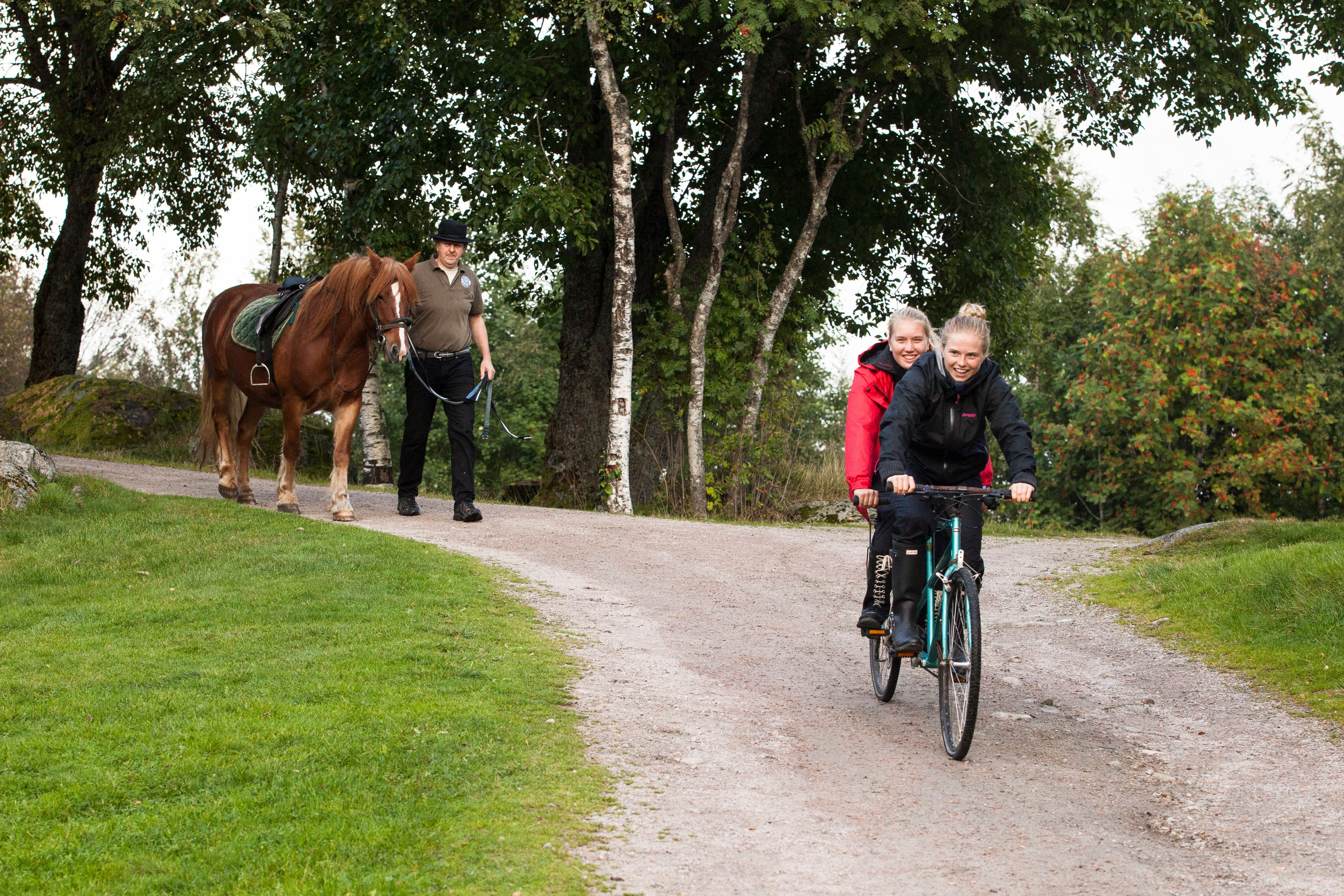 To smilende jenter sykler på en tandemsykkel, og en mann leier en brun hest med hvit bliss bak jentene.