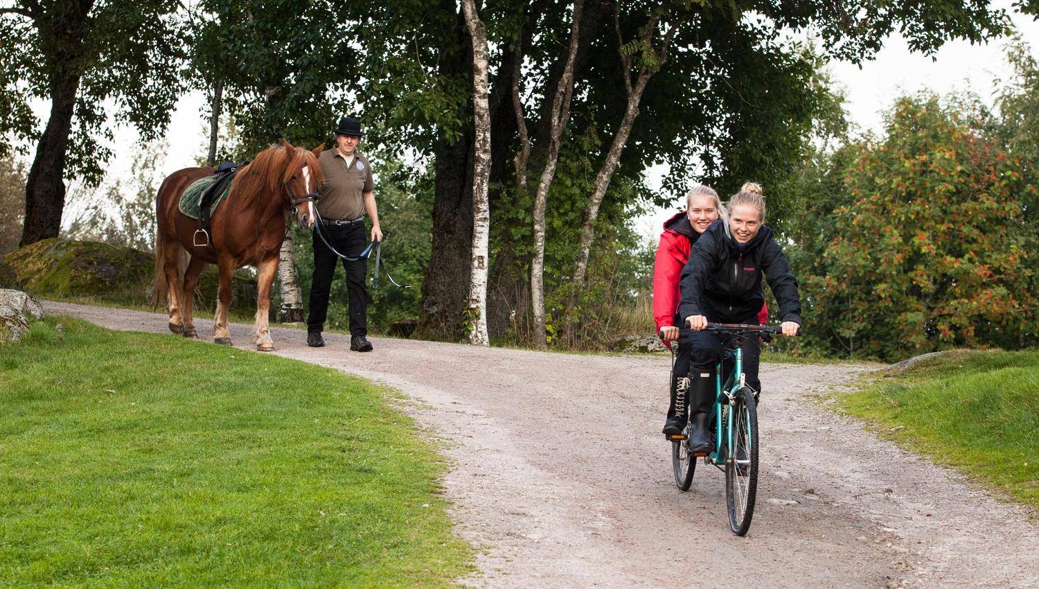 To smilende jenter sykler på en tandemsykkel, og en mann leier en brun hest med hvit bliss bak jentene.