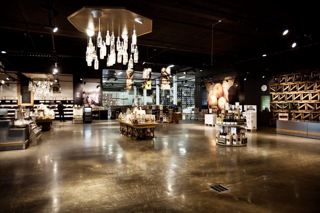 A modern store interior with shiny floors and bright lighting. Tables and shelves display bottles, jars, and local products. The ceiling lights are ma