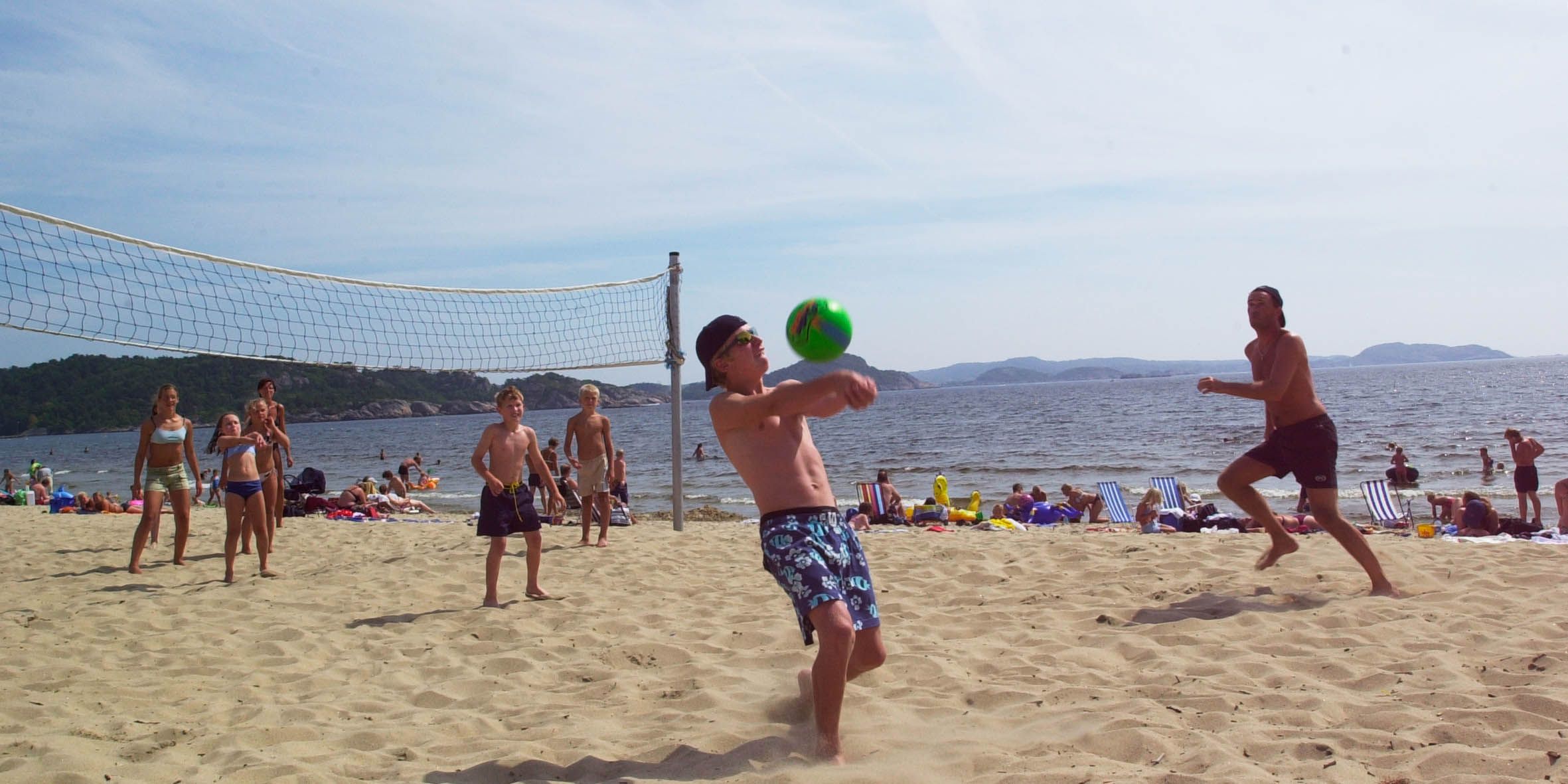 En gjeng som spiller strandvolleyball, der en fyr skal serve en grønn volleyball
