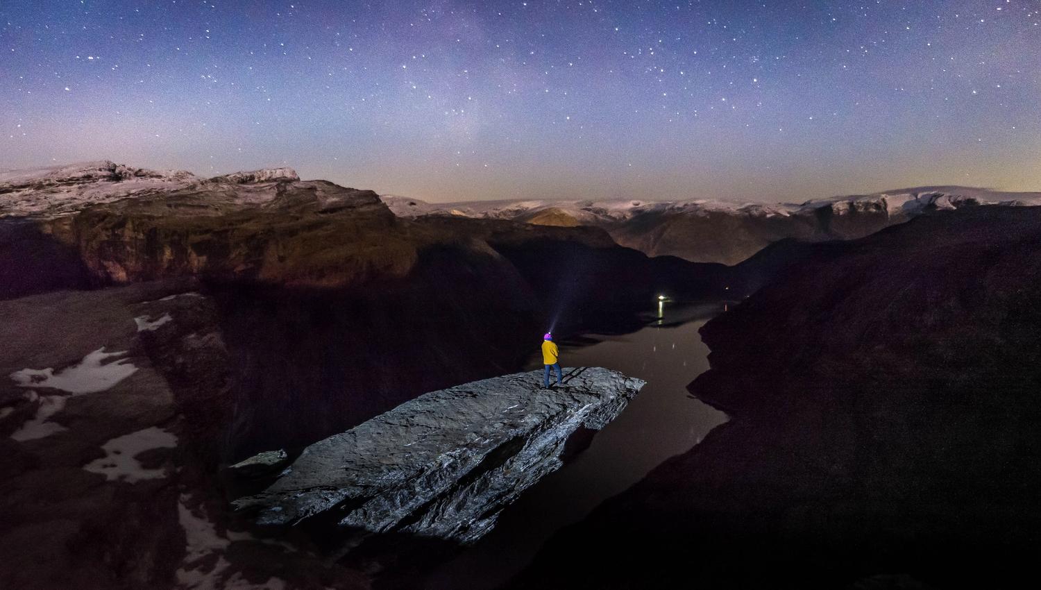 Hiker with headlamp standing on Trolltunga, overlooking the fjord under the night sky.