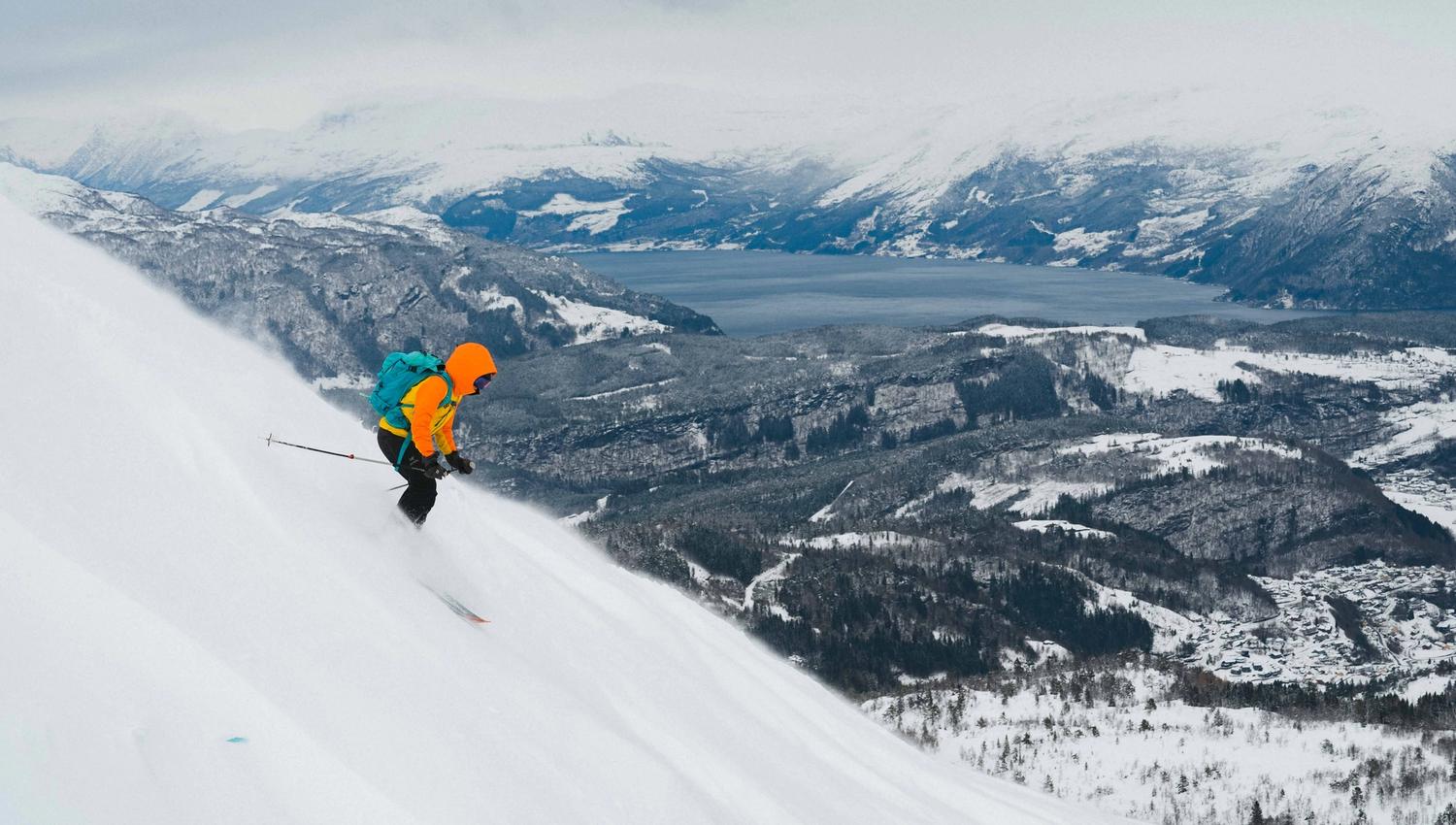 En person står på ski ned en bratt bakke med fjordutsikt.