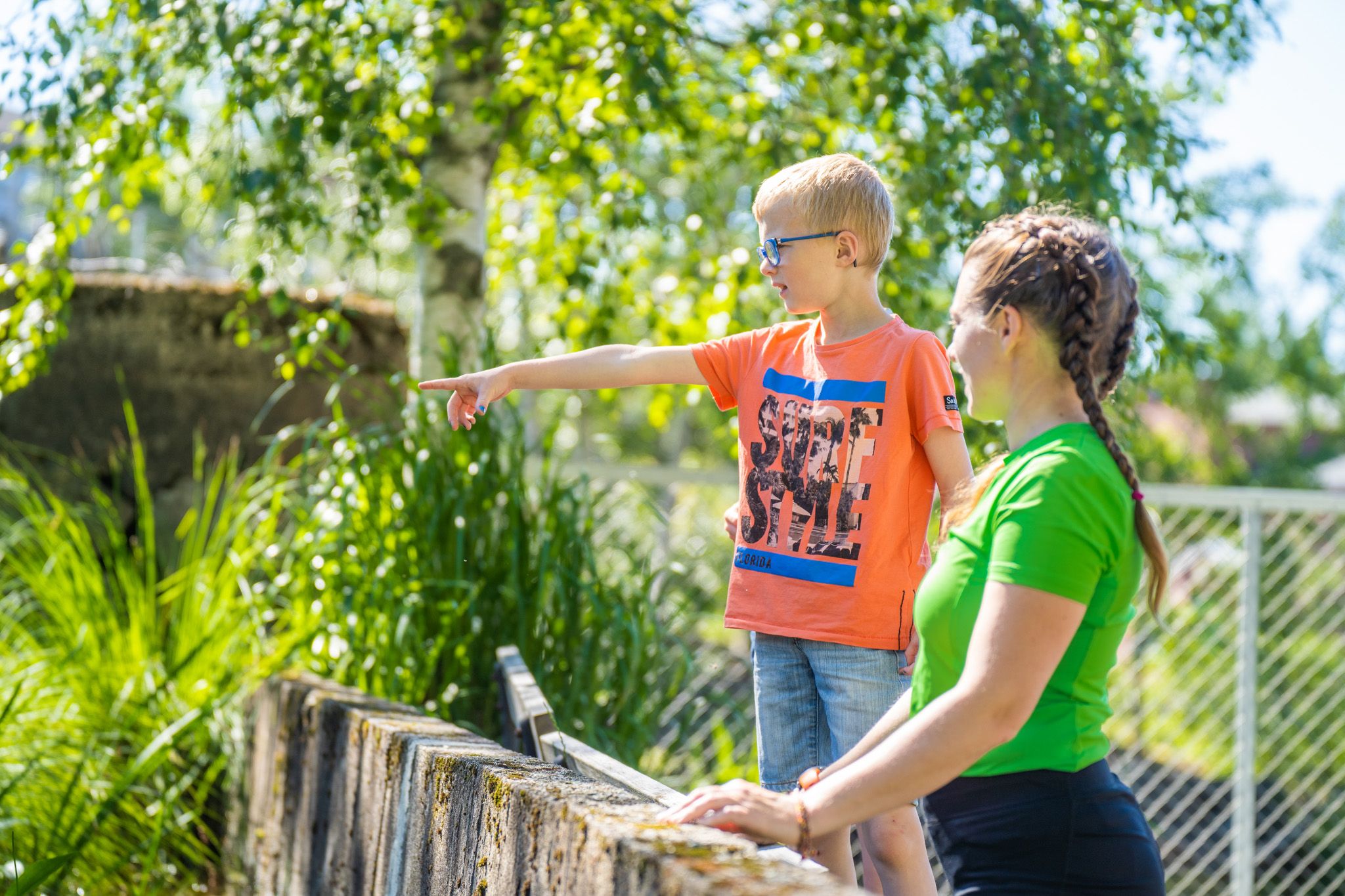 Bildet viser en jente og en gutt som står ute i sola. Gutten, som har på seg oransje t-skjorte og solbriller, peker på noe i det fjerne. Jenta, i grønn t-skjorte, står ved siden av og ser i samme retning. Bak dem er grønne trær og gress.