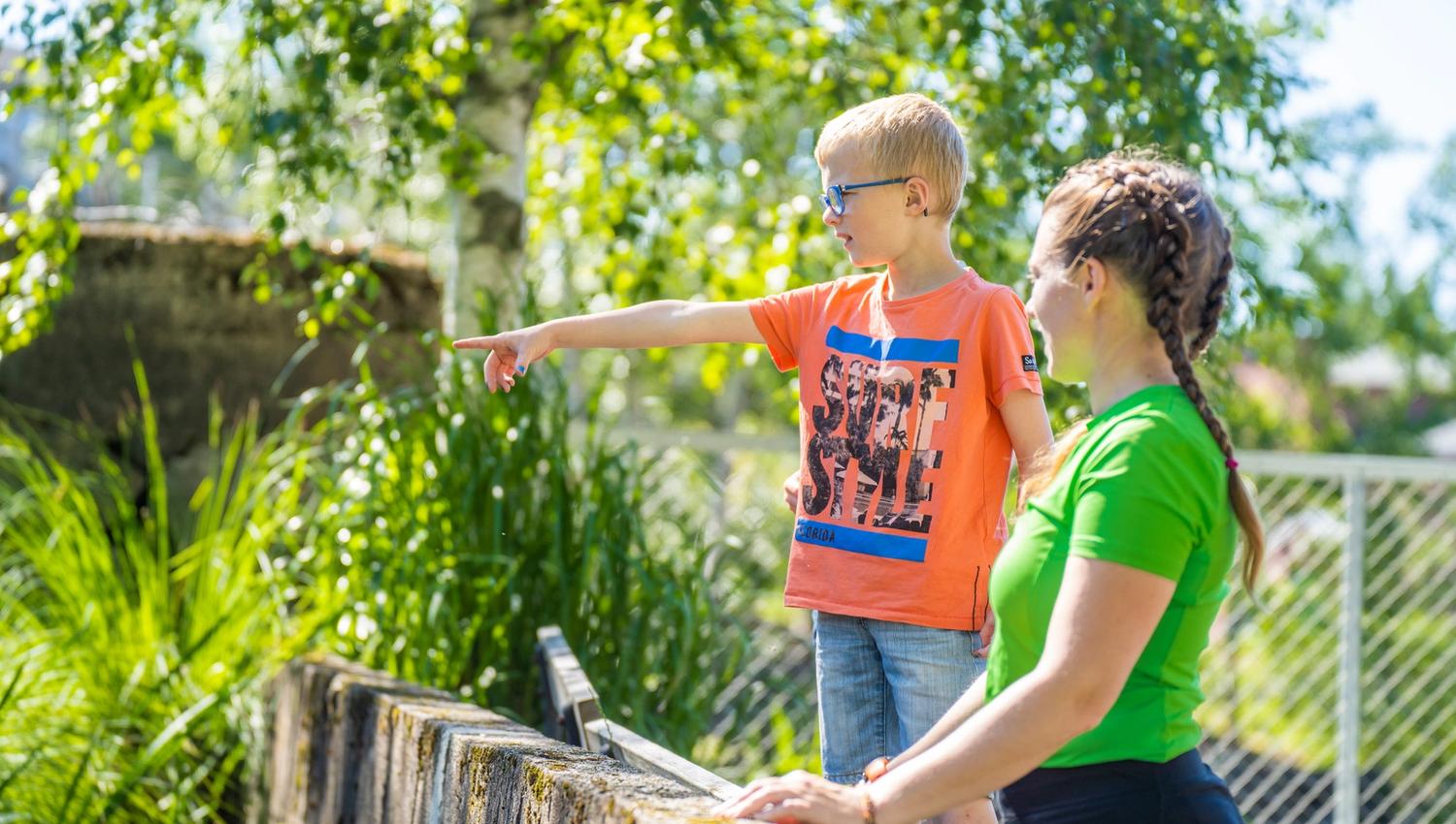 Bildet viser en jente og en gutt som står ute i sola. Gutten, som har på seg oransje t-skjorte og solbriller, peker på noe i det fjerne. Jenta, i grønn t-skjorte, står ved siden av og ser i samme retning. Bak dem er grønne trær og gress.