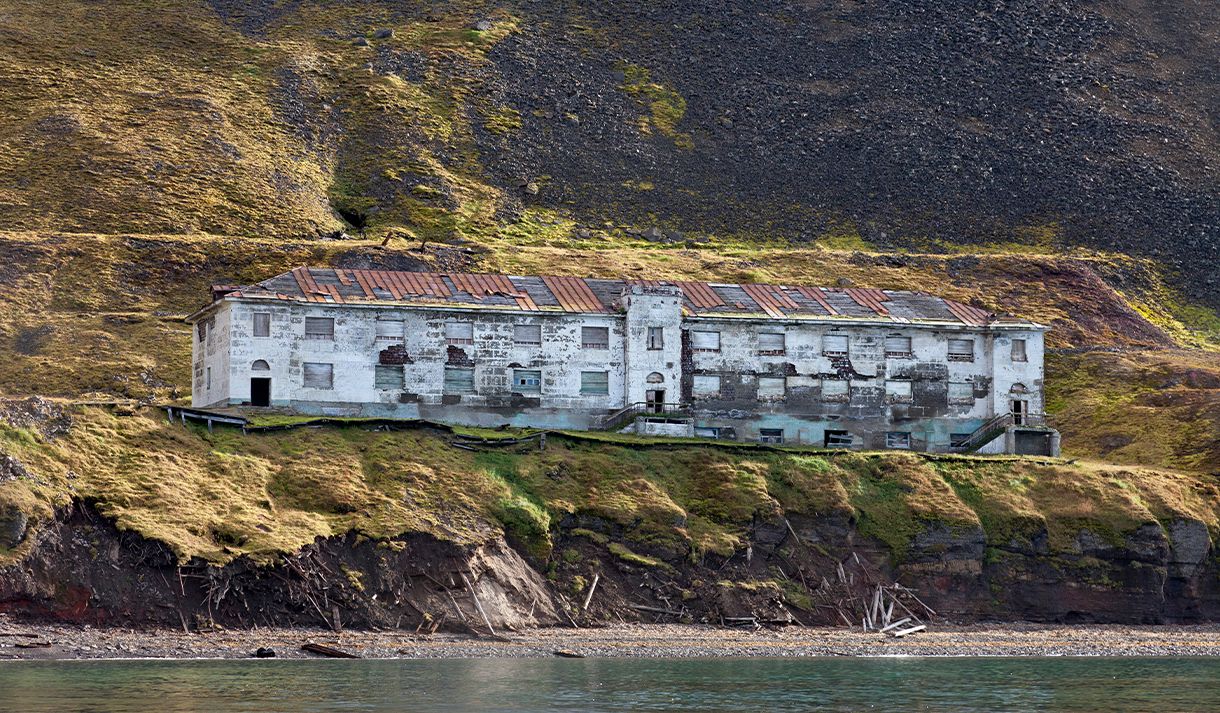 An abandoned building in the former mining community Grumant, with a beach and fjord in the foreground, and a mountain side in the background