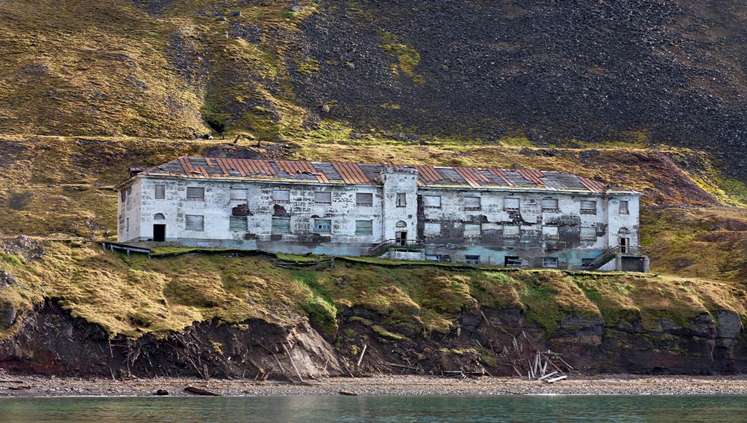 An abandoned building in the former mining community Grumant, with a beach and fjord in the foreground, and a mountain side in the background