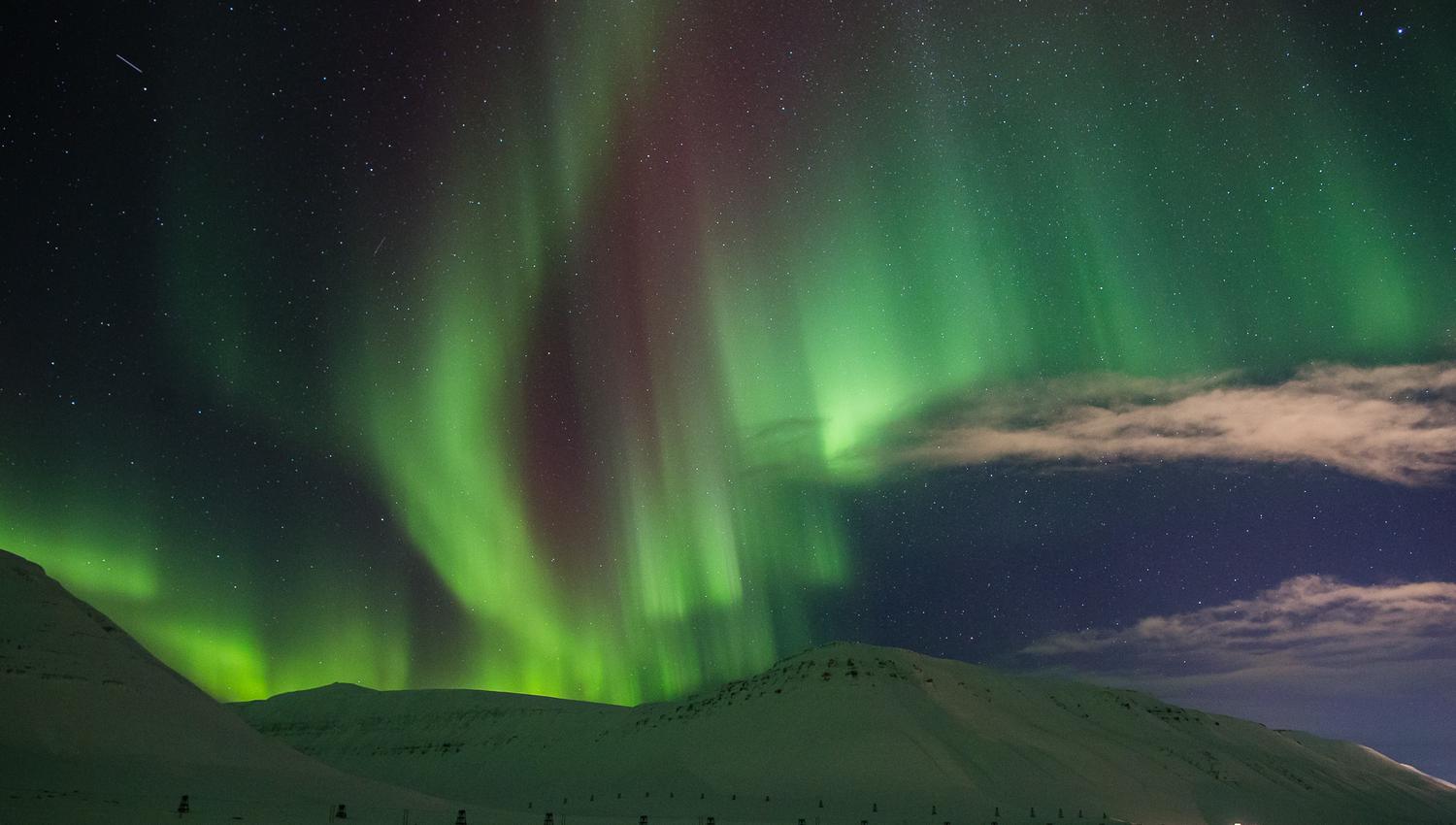 Et fjellandskap i forgrunnen med nordlys på himmelen i bakgrunnen