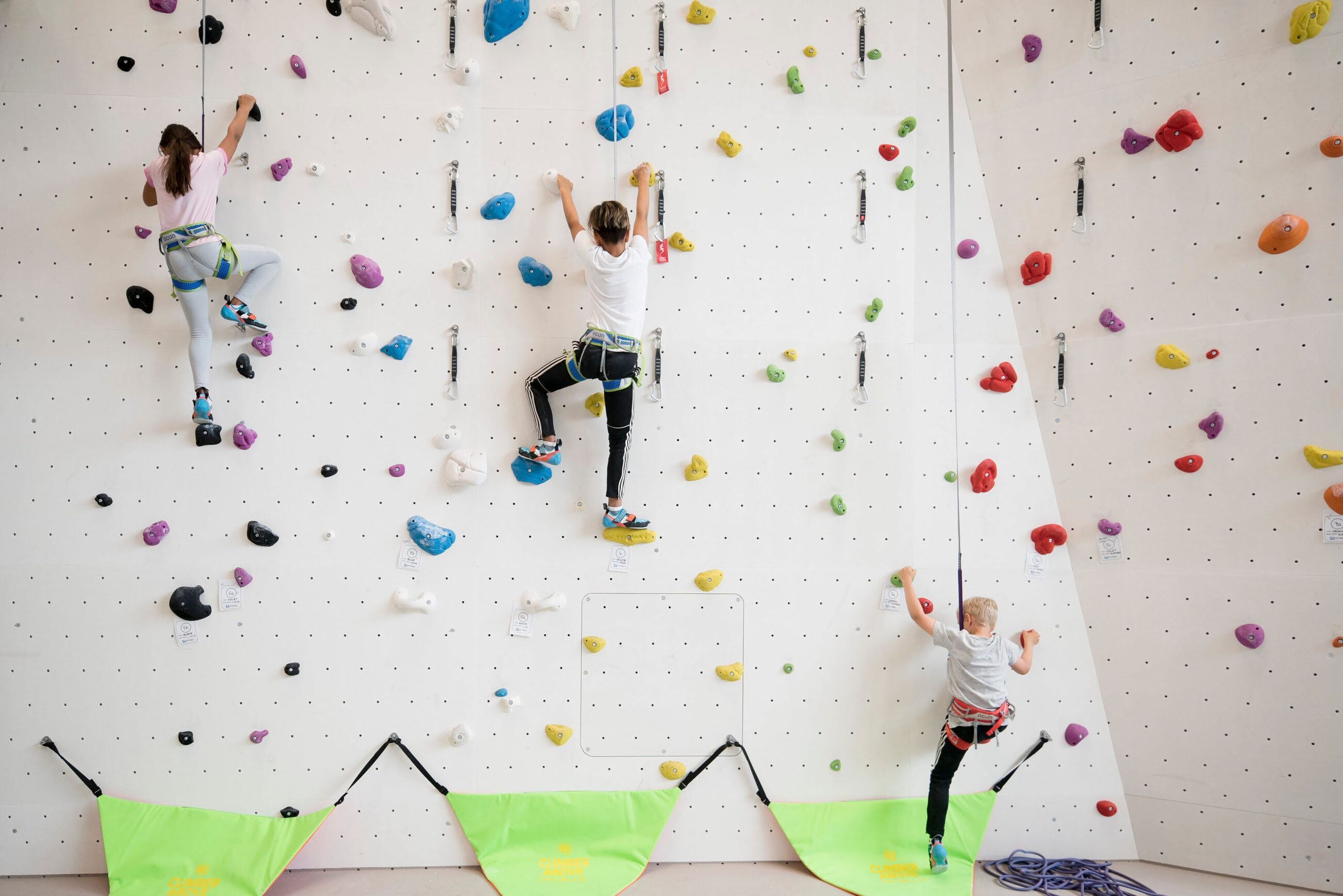 Three children climbing an indoor wall with colourful holds and safety equipment.