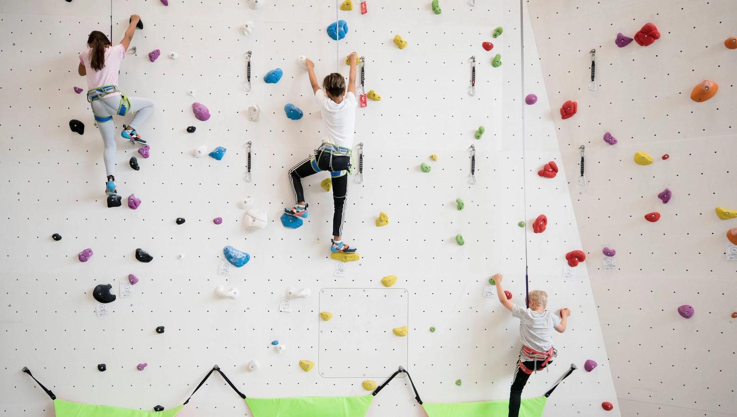 Three children climbing an indoor wall with colourful holds and safety equipment.