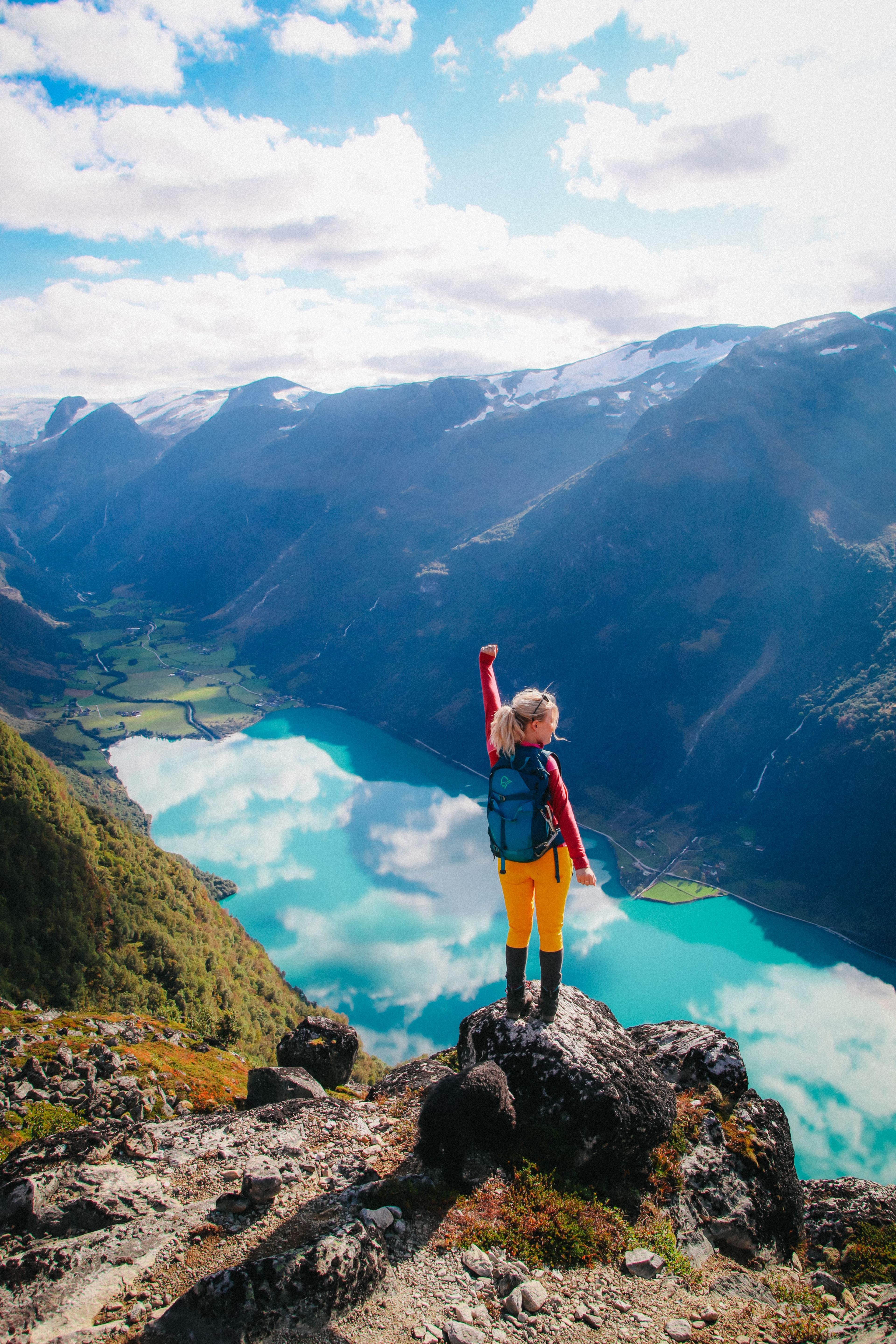 A hiker with a backpack stands on a rocky cliff above a turquoise lake, with mountains and clouds in the distance..