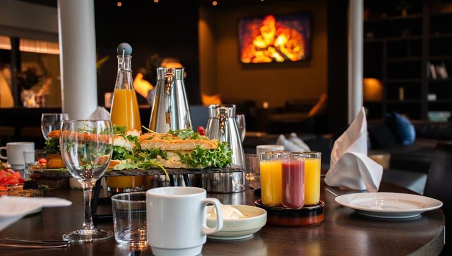 Bord med frokostservering, juice, kaffe og glass, i en lun og stemningsfull lounge.