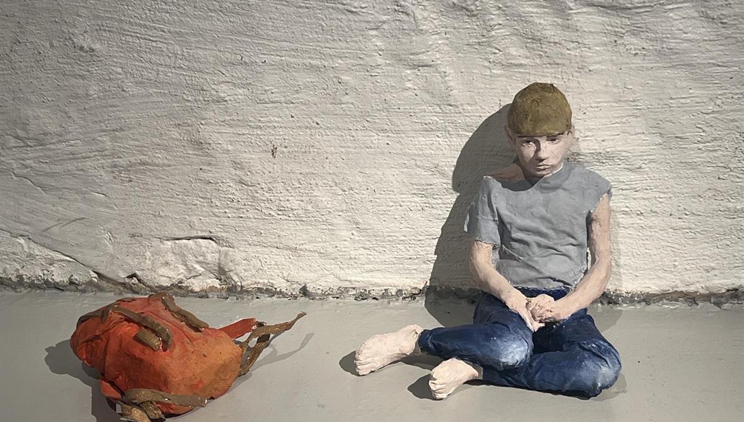 Figur sitter på gulvet ved en hvit murvegg med jakke lagt ved siden av.