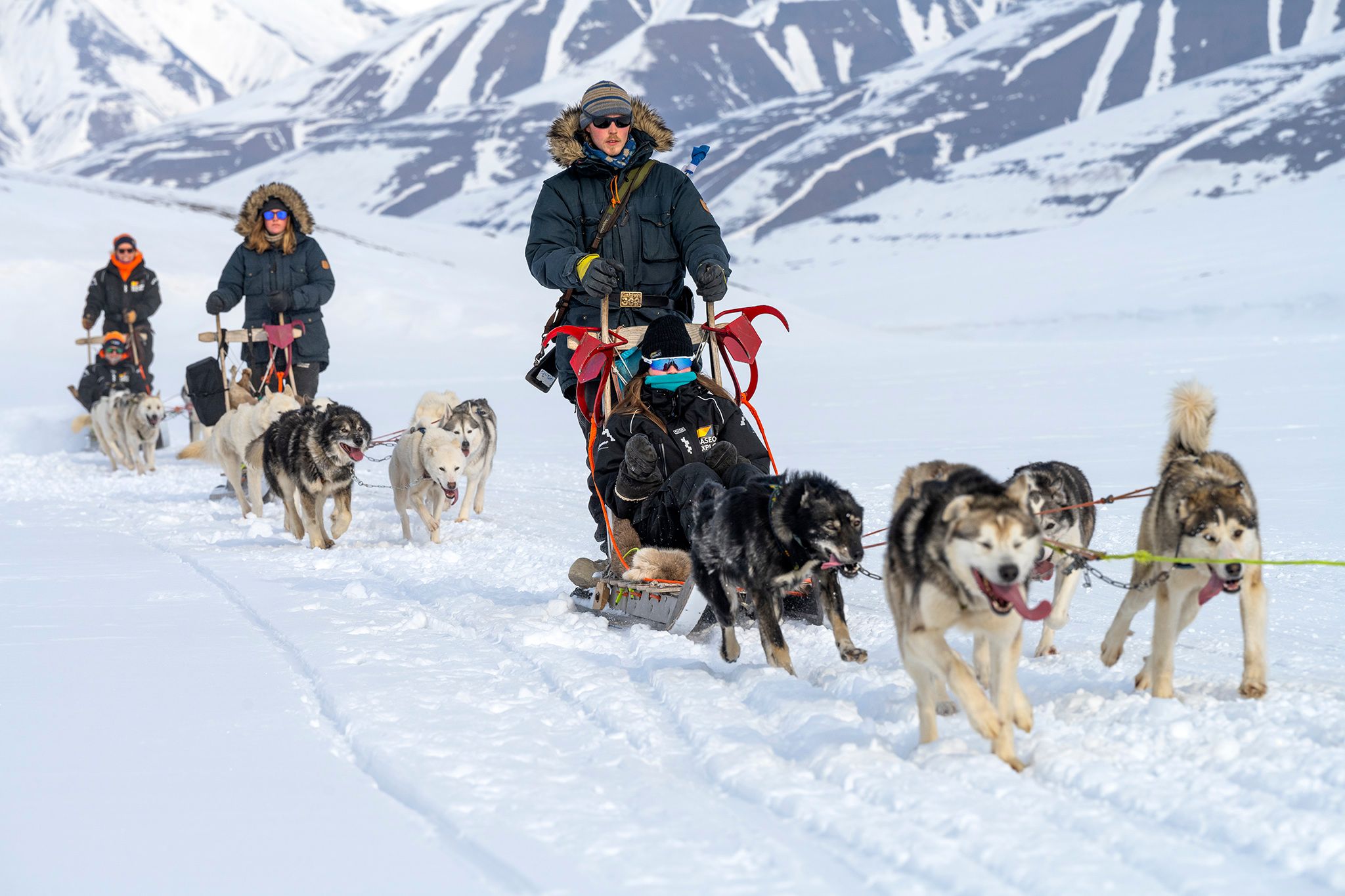 A guide driving a dog sled with a guest sitting on it. In the background, two more sleds are following along.
