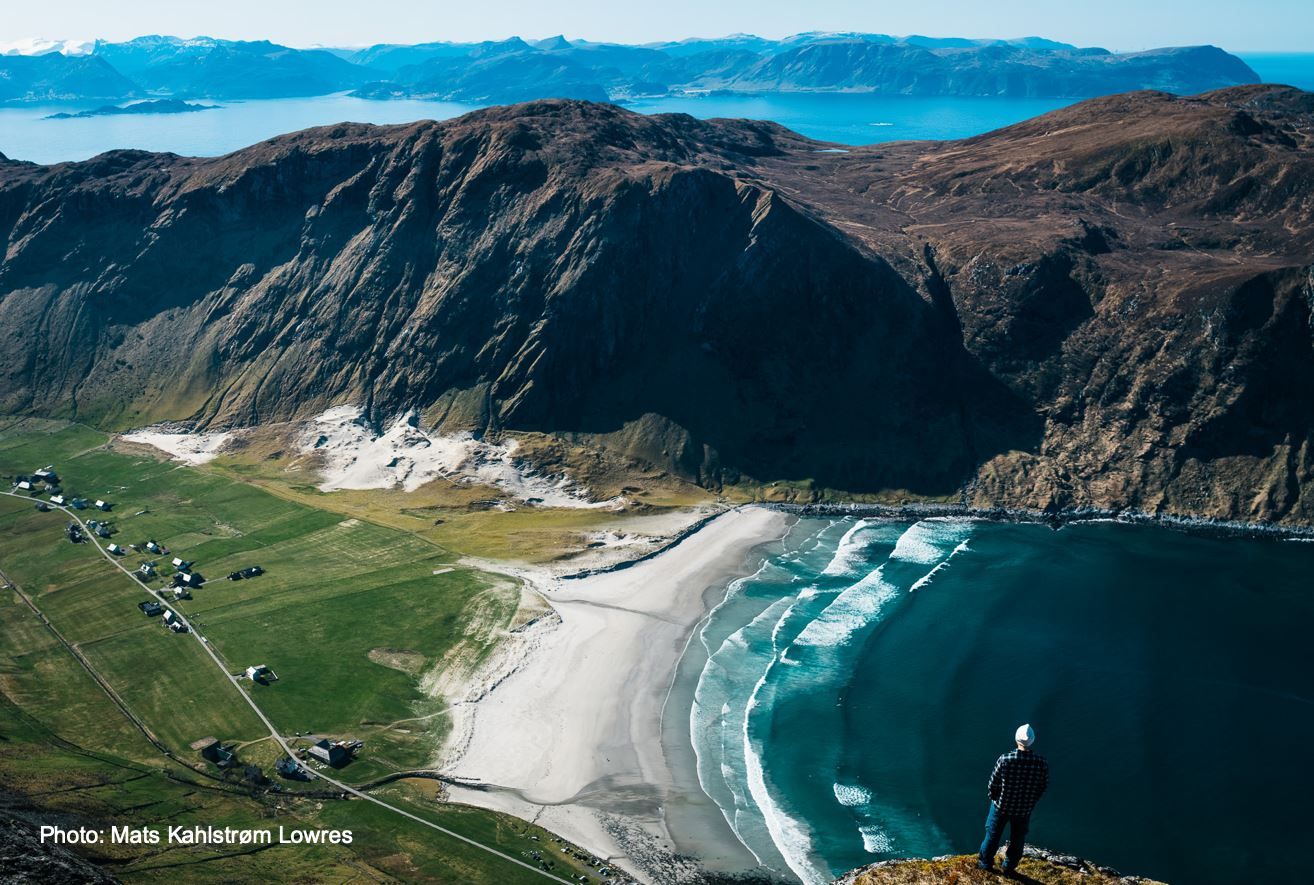 Person stands on a rocky cliff above a curved sandy beach, green fields, and rolling ocean waves beneath mountains..
