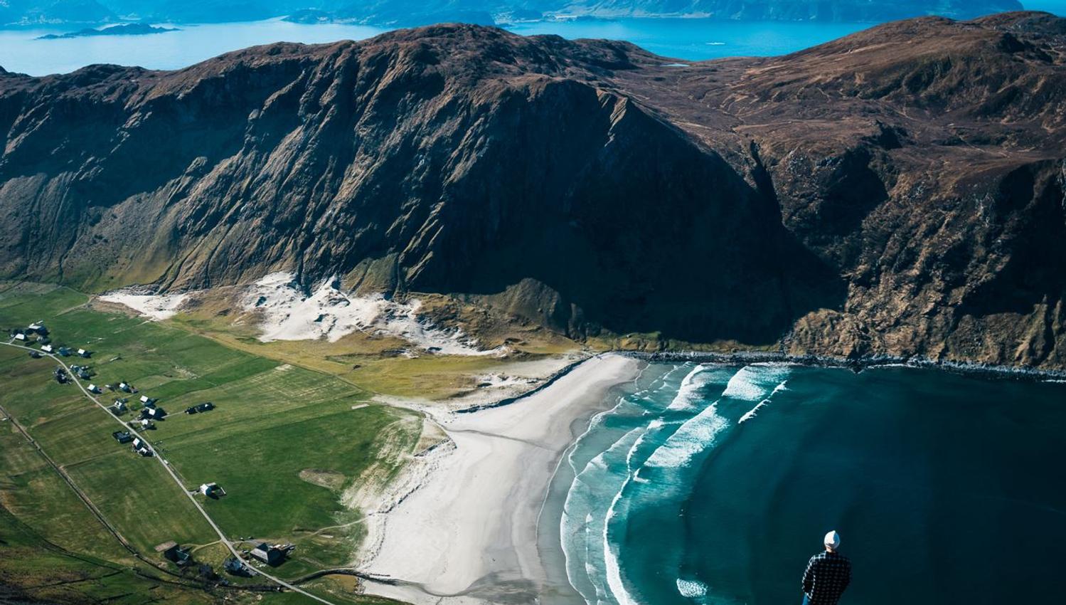 Person stands on a rocky cliff above a curved sandy beach, green fields, and rolling ocean waves beneath mountains..