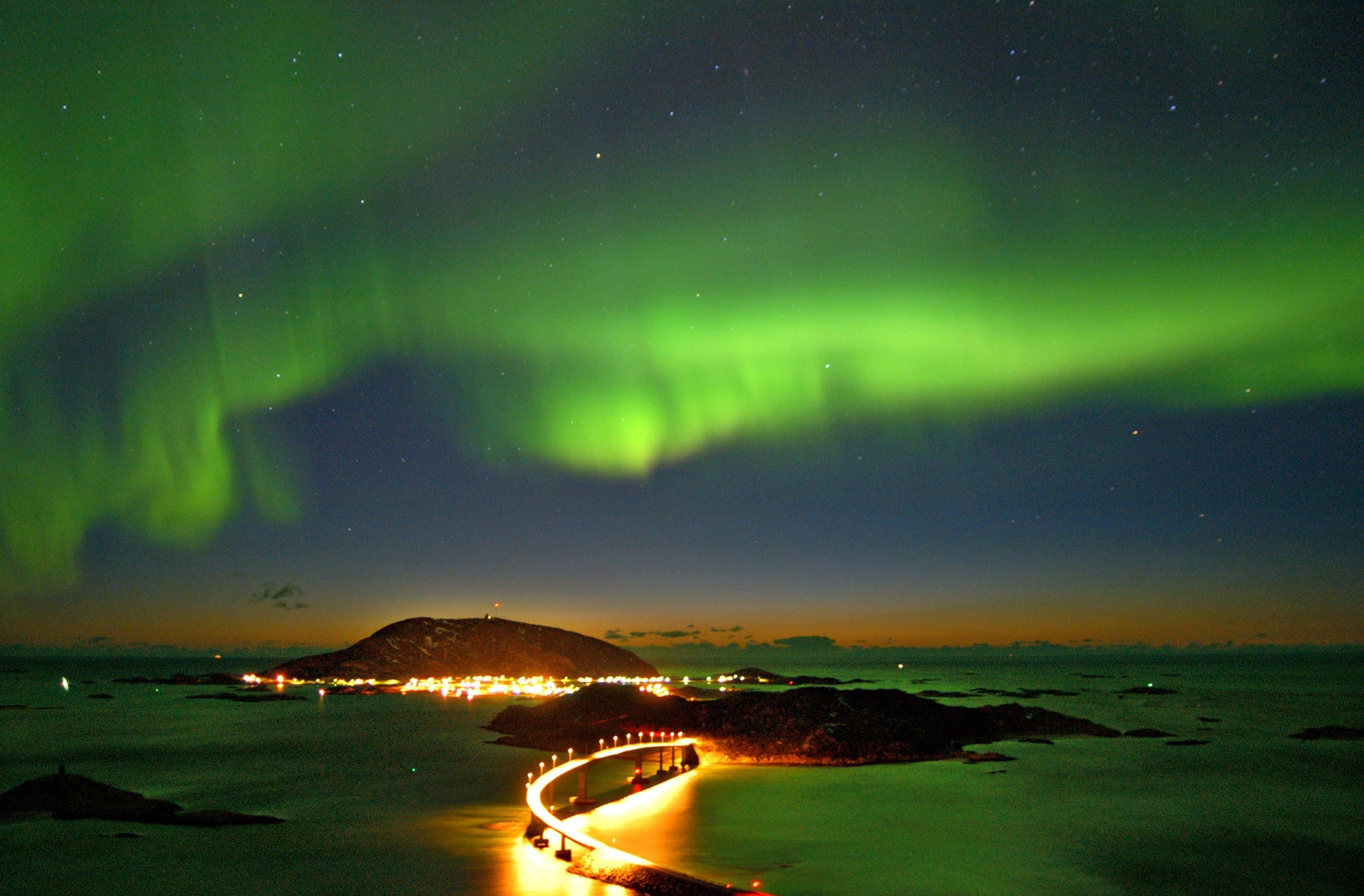 Green-hued northern lights in the sky, with a lit road stretching across dark water.