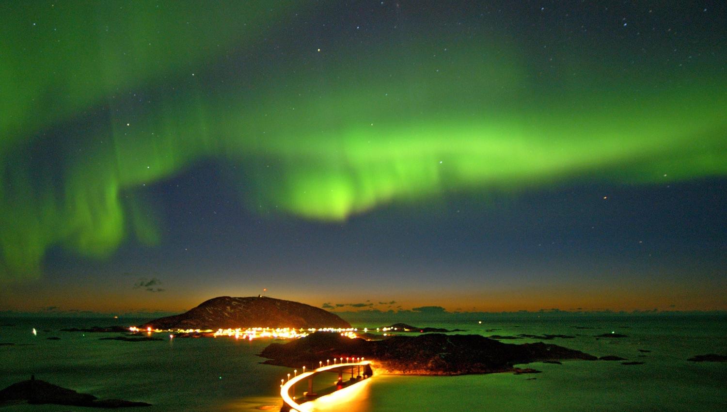 Green-hued northern lights in the sky, with a lit road stretching across dark water.