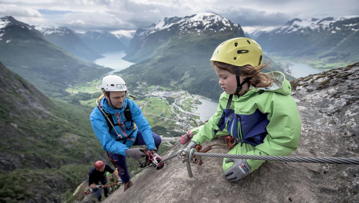 To klatrarar med hjelm og sele på ei bratt fjellhylle, festa i stålvaier med fjord og fjell i bakgrunnen..