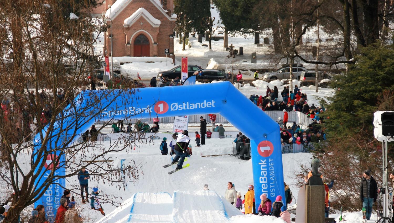 En skiløper hopper over et snødekt hopp, med publikum langs løypa og en blå oppblåsbar bue i bakgrunnen..